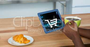 Cropped image of person holding digital tablet with shopping cart icon on screen while having coffee
