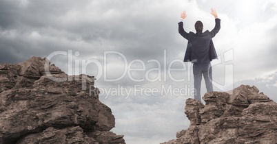 Business man hands in air on rocks against clouds with flare