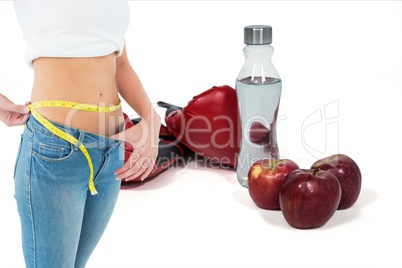 Midsection of woman in loose jeans measuring waist with apples and boxing gloves with bottle in back