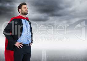 Business man superhero with hands on hips against road and grey sky with flare