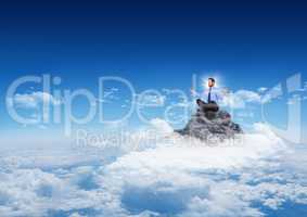 Business man with flare meditating on mountain peak in the clouds with blue sky