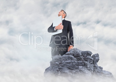 Business man with laptop looking up on rock in clouds