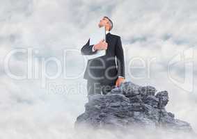 Business man with laptop looking up on rock in clouds