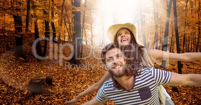 Happy man with arms outstretched carrying woman on back in forest during autumn