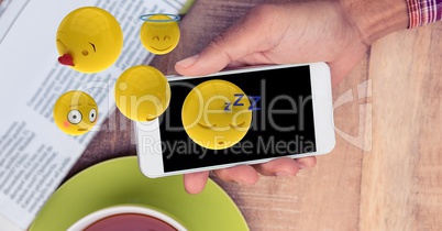 Digitally generated image of emojis flying over hand using smart phone at table