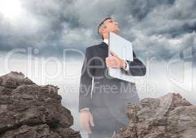 Business man with laptop and flare looking up behind rocks