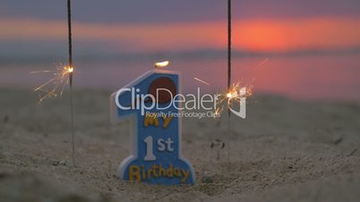 Slow motion view of candle and two sparklers standing in the sand on beach against blurred sunset