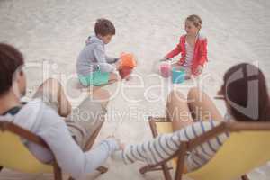 Siblings playing by parents relaxing on launge chairs at beach