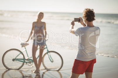 Man photographing woman standing by bicycle at beach