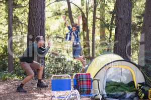 Father photographing playful boy jumping at campsite