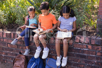 Full length of friends reading books while sitting on retaining wall