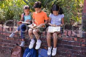 Full length of friends reading books while sitting on retaining wall