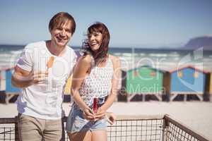 Happy couple eating ice cream standing by railing at beach