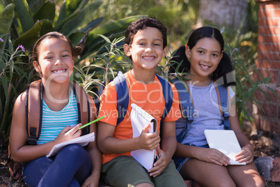 Smiling friends with books and digital tablets sitting on retaining wall