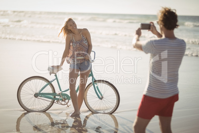 Smiling woman standing by bicycle while man photographing her at beach