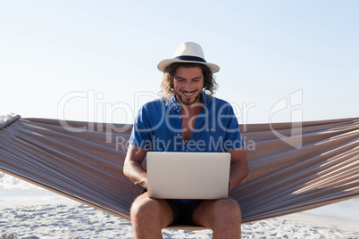 Smiling Man using laptop while sitting on hammock at beach