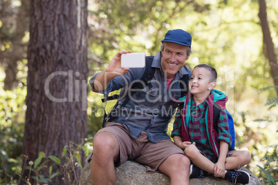 Happy father sitting with boy on rock taking selfie