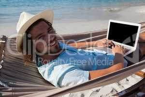 Smiling man using laptop while relaxing on hammock in beach