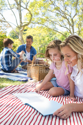 Mother and daughter reading book while father and son playing with football in park