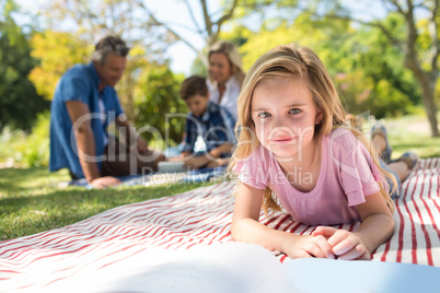 Girl lying on blanket and reading book while family sitting in background