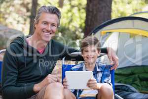 Happy father and son with digital tablet by tent in forest