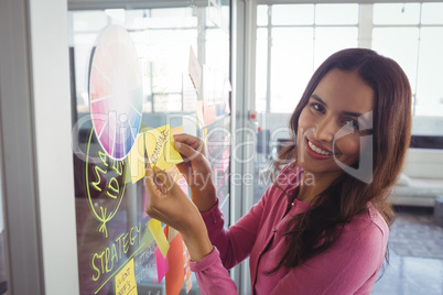 Smiling female designer holding adhesive note on glass in creative office