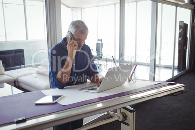 Businessman talking on phone while using laptop in office