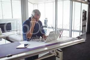 Businessman talking on phone while using laptop in office