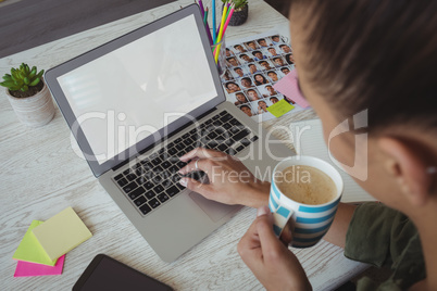 Female photo editor holding coffee cup while using laptop in office