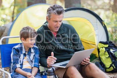 Father and son using laptop by tent in forest
