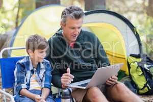 Father and son using laptop by tent in forest