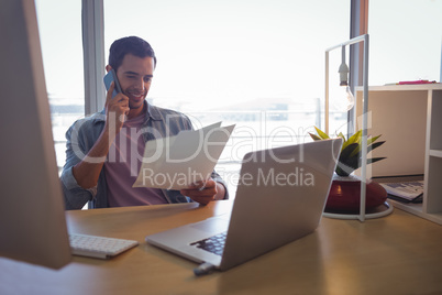 Businessman holding photographs while talking on mobile phone at office