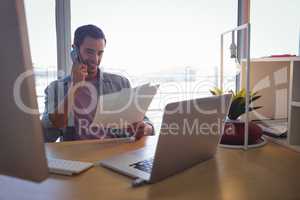 Businessman holding photographs while talking on mobile phone at office