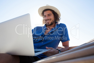 Smiling man using laptop while relaxing on hammock at beach