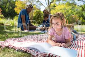 Girl lying on blanket and reading book while family sitting in background