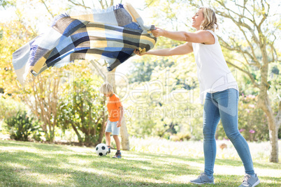 Mother spreading the picnic blanket while son playing football in background