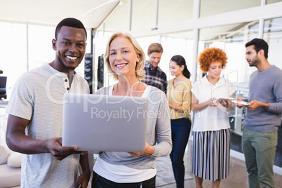 Portrait of smiling business people holding laptop with colleagues in background