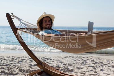 Smiling man using laptop while relaxing on hammock in beach