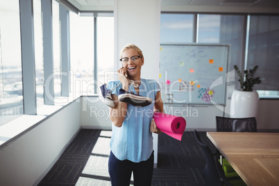 Smiling executive talking on mobile phone while holding exercise mat and shoes