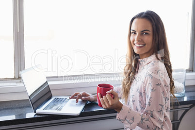 Portrait of smiling businesswoman holding coffee cup while working on laptop