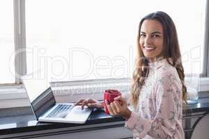 Portrait of smiling businesswoman holding coffee cup while working on laptop