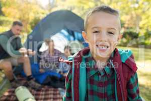 Boy smiling at camera while family sitting at tent in background