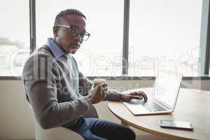 Portrait of confident executive having coffee while using laptop