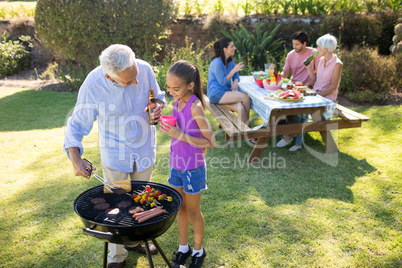 Grandfather and granddaughter preparing barbecue while family having meal