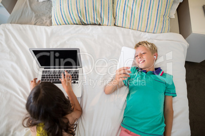 Siblings using mobile phone and laptop on bed