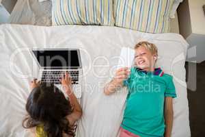 Siblings using mobile phone and laptop on bed