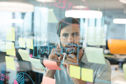 Businessman using mobile phone while looking at plans written on glass