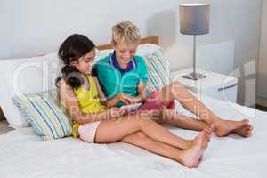 Smiling siblings using digital tablet on bed in bedroom