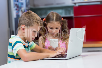 Siblings using laptop and mobile phone in kitchen