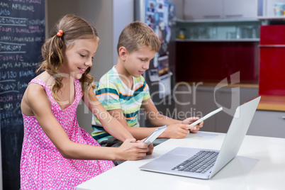 Siblings using digital tablet and mobile phone in kitchen